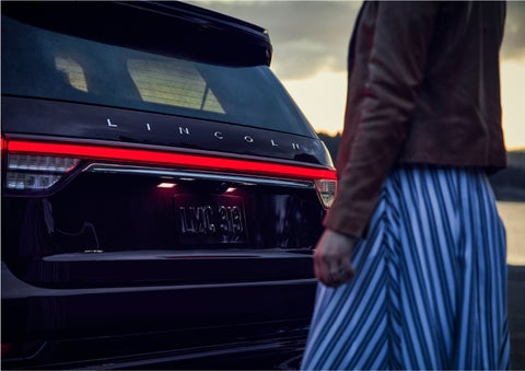 A person is shown near the rear of a 2023 Lincoln Aviator® SUV as the Lincoln Embrace illuminates the rear lights | White's Canyon Motors - Lincoln in Spearfish SD