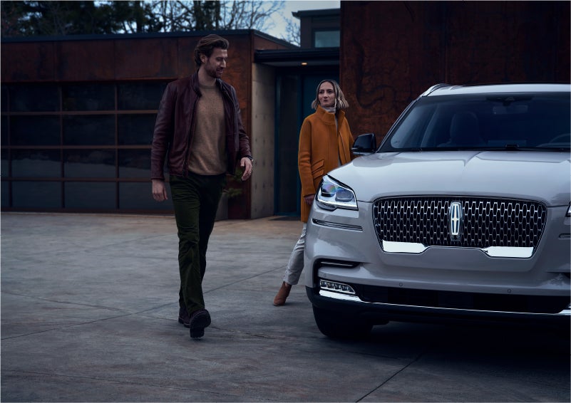 A man and a woman approaching a 2023 Lincoln Aviator® SUV, which illuminates certain lights when they are close | White's Canyon Motors - Lincoln in Spearfish SD